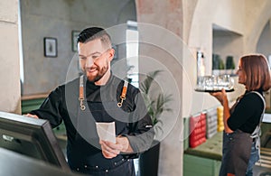 Stylish bearded smiling waiter dressed black uniform using point of sale order terminal system touch Screen and waitress woman