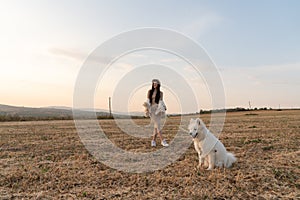 Stylish beautiful brunette girl in beige dress, coat and hat stands among the field with samoed dog on a leash