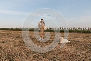 Stylish beautiful brunette girl in beige dress, coat and hat stands among the field with samoed dog on a leash