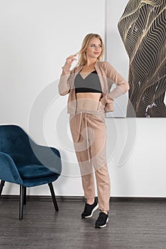 Stylish beautiful young blond woman in a biege tracksuit poses near a white wall in the room