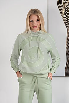 Stylish beautiful young blond woman in a green tracksuit poses near a white wall in the room.