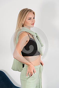Stylish beautiful young blond woman in a green tracksuit poses near a white wall in the room