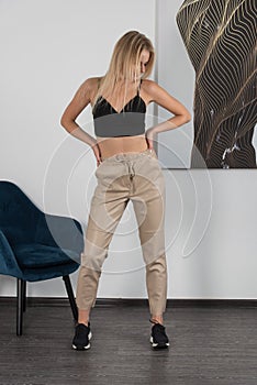 Stylish beautiful young blond woman in a tight biege pants, black top and white shirt near a white wall in the room. Attractive