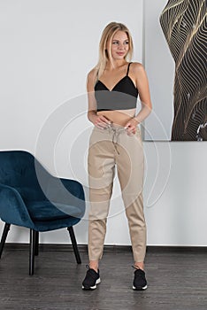 Stylish beautiful young blond woman in a tight biege pants, black top and white shirt near a white wall in the room. Attractive