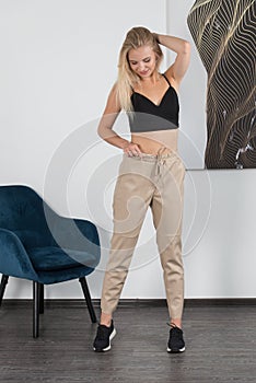 Stylish beautiful young blond woman in a tight biege pants, black top and white shirt near a white wall in the room. Attractive