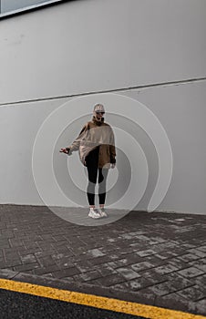 Stylish beautiful young woman in a fashionable leather jacket with black leggings and sneakers stands near a gray urban wall on