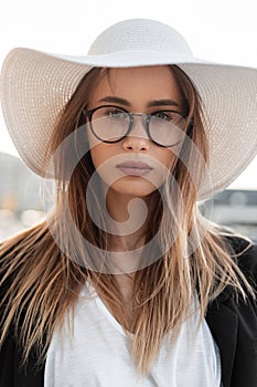 Stylish beautiful young woman in glasses in a fashionable hat with a black jacket and white T-shirt on the street. Young