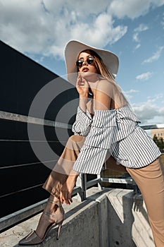 Stylish beautiful young woman model in trendy sunglasses in a fashion summer hat with a top and trousers puts on shoes in the city