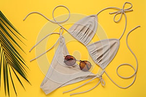 Stylish beige bikini, sunglasses and leaf on yellow background, flat lay