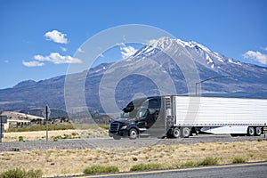 Stylish black big rig bonnet semi truck tractor transporting cargo in refrigerator semi trailer running on the highway road past