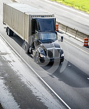 Stylish black big rig day cab semi truck with roof spoiler transporting cargo in container running on the straight divided highway