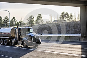 Stylish black day cab big rig semi truck transporting fuel in tank semi trailer running on the sunny road under the bridge