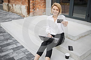 Stylish blond businesswoman doing online purchases while sitting outside near office building.