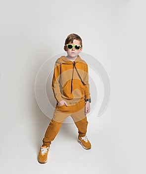 Stylish blond kid boy in sunglasses Frolic excited child in yellow ginger sportswear hoodie and pants stands with his