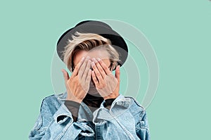 Stylish blond woman closes eyes with her hands in jeans jacket and hat on green background