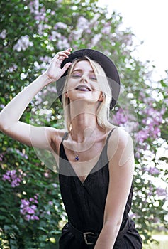 Stylish blonde with a black hat on a background of blooming lilac, a young white woman smiling