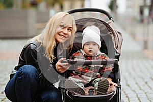 Stylish blonde mother walking with her little daughter in a stroller outdoors.