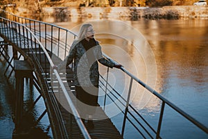 Stylish blonde stands on narrow bridge over autumn pond