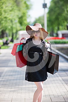 Stylish blonde woman in black dress, sunglasses and hat with shopping bags walking in the city