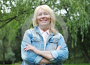 Stylish blonde woman in a denim jacket in a summer park. An elderly happy lady pensioner stands with her arms crossed.