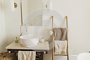 Stylish boho bathroom design. Modern ceramic sink with golden faucet on rustic stand with soap and ladder with towel on background