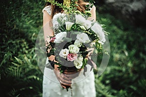 Stylish boho bride holding rustic bouquet of amazing flowers and herbs on background of rocks in mountains