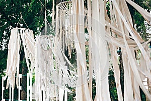 Stylish boho decor on trees. Modern bohemian decoration of white macrame and ribbons, hanging on branches in summer park. Wedding