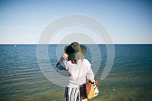 Stylish boho girl in hat looking at sea in sunny evening light from sandy cliff, back view. Happy young hipster woman relaxing on