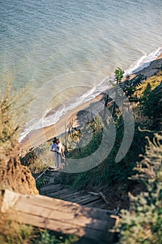 Stylish boho girl in hat standing at sandy cliff with grass at sea in sunny light. Happy young hipster woman exploring tropical