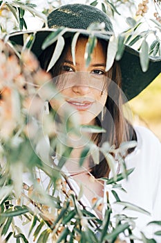 Stylish boho girl with modern earrings posing among green olive branches in soft evening light. Happy young fashionable woman