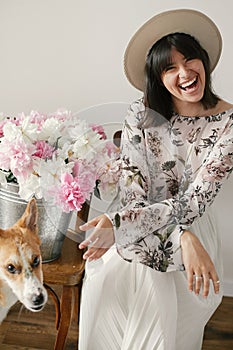 Stylish boho girl sitting at metal bucket with peonies on rustic wooden chair and playing with cute golden dog.Beautiful hipster