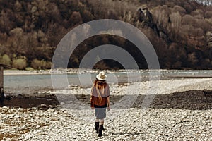 stylish boho traveler woman in hat back view, fringe poncho posing near water river beach in mountains, gypsy hipster girl.