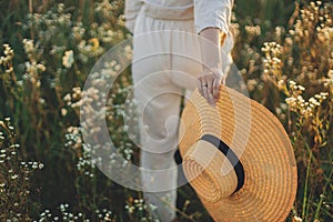 Stylish boho woman with straw hat in hand close up among wildflowers in sunset light. Atmospheric moment. Summer delight and