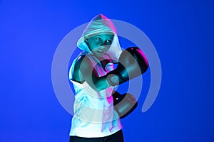 Stylish boy, beginner boxer training isolated over blue background in neon light. Concept of sport, fashion, studying