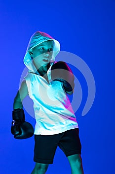 Stylish boy, beginner boxer training isolated over blue background in neon light. Concept of sport, fashion, studying