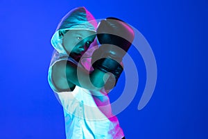 Stylish boy, beginner boxer training isolated over blue background in neon light. Concept of sport, fashion, studying