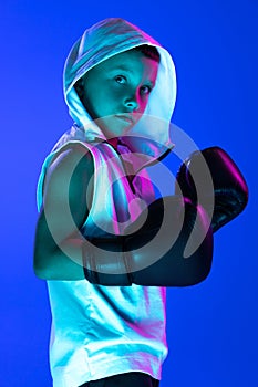 Stylish boy, beginner boxer training isolated over blue background in neon light. Concept of sport, fashion, studying