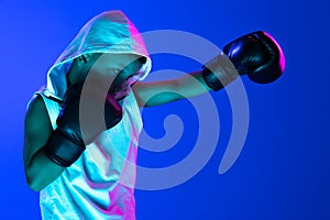 Stylish boy, beginner boxer training isolated over blue background in neon light. Concept of sport, fashion, studying