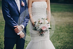 Stylish bride with bouquet and groom standing in wedding aisle with rose petals on grass during matrimony, cropped view .
