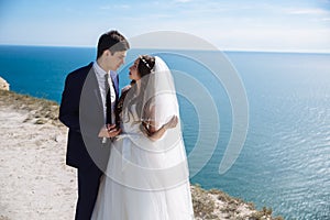 Stylish bride and elegant groom look at each other with love at photo shooting on cliff with ocean view