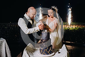 Stylish bride and groom cut the wedding cake in the evening on the street