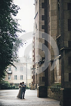 Stylish bride and groom dancing on background of old church in rainy street. Provence wedding. Beautiful emotional wedding couple