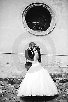 Stylish bride and groom gently hugging in european city street. Gorgeous wedding couple of newlyweds embracing near ancient