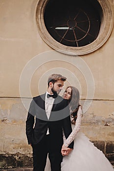 Stylish bride and groom gently hugging in european city street. Gorgeous wedding couple of newlyweds embracing near ancient