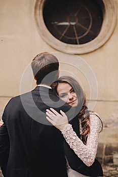 Stylish bride and groom gently hugging in european city street. Gorgeous wedding couple of newlyweds embracing near ancient