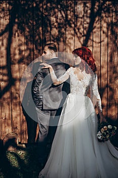 Stylish bride and groom posing in sunlight on background of wooden wall in country. rustic wedding concept in barn, space for text