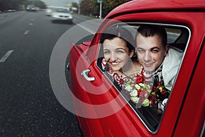 Stylish bride and happy groom looking out of driving  retro car, having fun and kissing