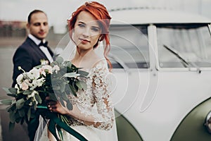 Stylish bride holding modern bouquet and groom looking at her near retro car. luxury wedding couple newlyweds posing. space for