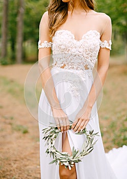 Stylish bride in a lace embroided dress with a cutout on the leg holding a wreath of olive branches