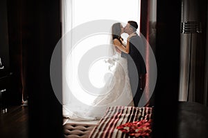 Bridegroom and bride in long dress are kissing near the window